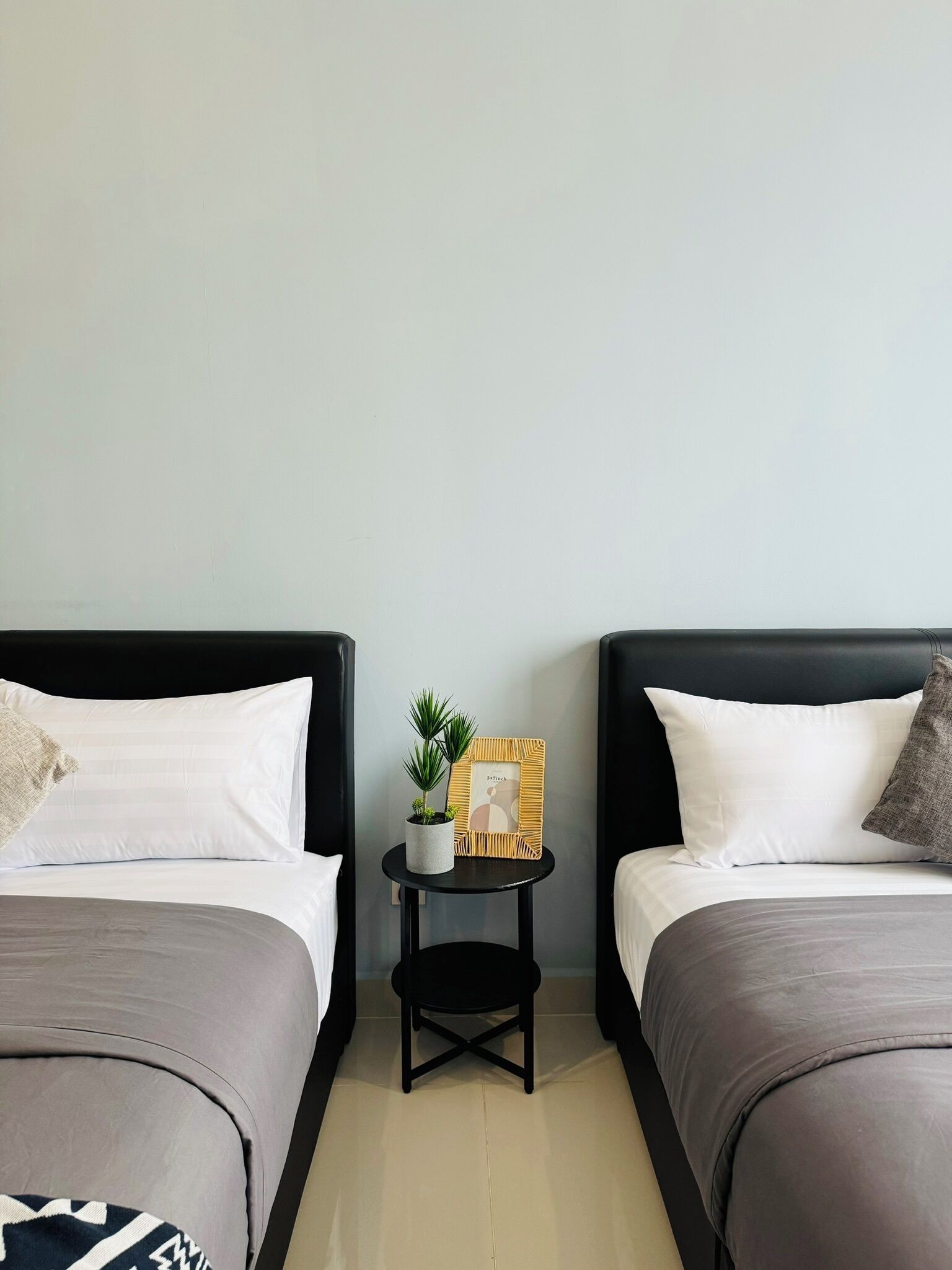 A hotel room with two beds, a small table with a plant and picture frame, and a plain wall.