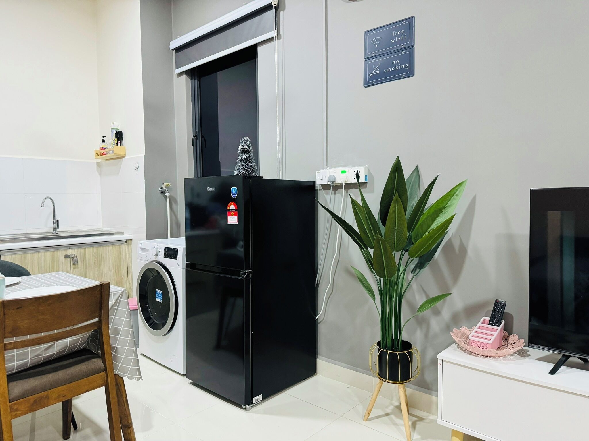 Une cuisine compacte équipée d’un lave-linge, d’un réfrigérateur noir, d’une plante en pot et d’un téléviseur posé sur un meuble.