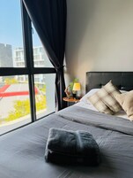 A modern hotel room with a large window, a bed with grey bedding, a black headboard, and a small bedside table with a lamp and decorative items.