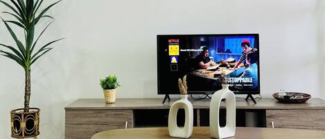Una sala de estar con televisor, una mesa de centro, una planta en maceta y cuadros en la pared.