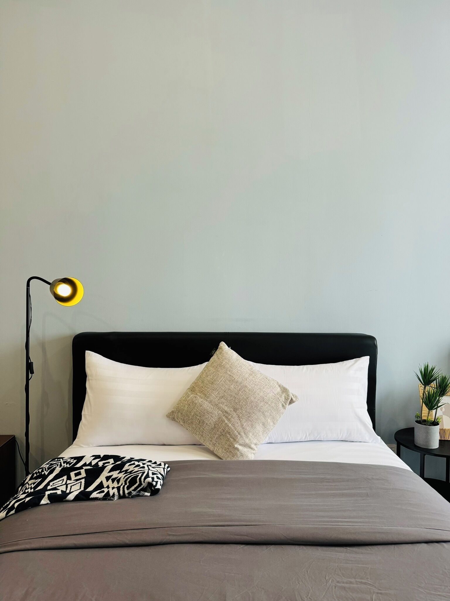 A bed with a black headboard, white and beige pillows, a grey blanket, and a black and white throw. There is a small side table with a plant and a lamp with a yellow shade.