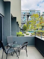 A balcony with a glass table and chairs, a view of a modern building, and a small garden area.