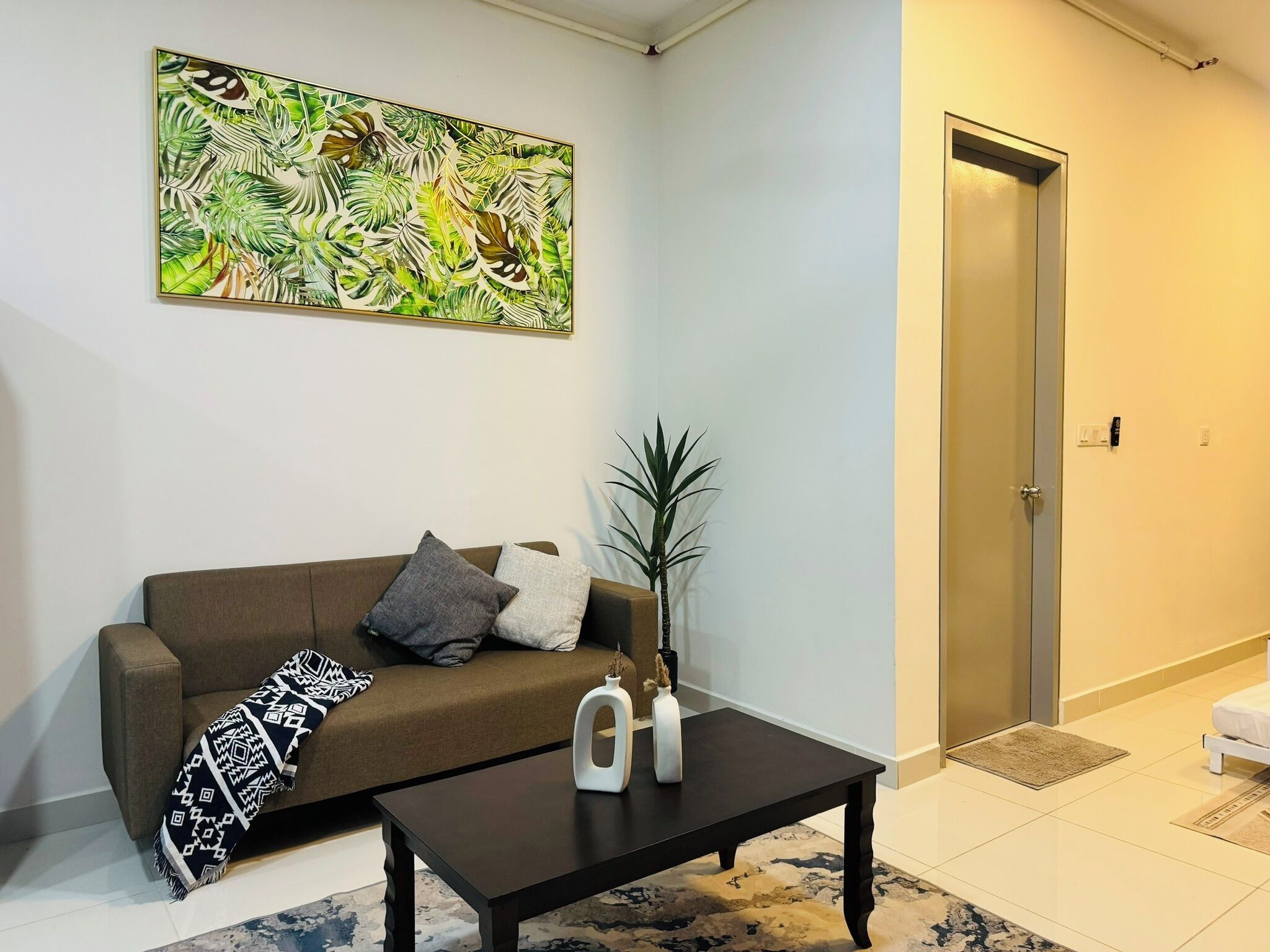 A living room with a brown sofa, two pillows, a patterned throw, a coffee table with decorative items, a large abstract painting, and a door.