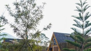 A small, triangular wooden cabin with a stone wall and a wooden door, surrounded by a bamboo fence and lush greenery. - RoomQuest Kid Tung Khao (Pak Chong)