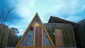 A triangular wooden structure with a stone facade and a wooden door, surrounded by bamboo fencing. - RoomQuest Kid Tung Khao (Pak Chong)