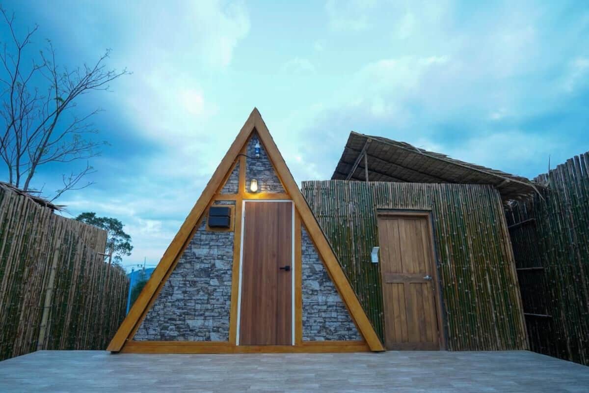 Una construcció de fusta triangular amb façana de pedra i porta de fusta, envoltada per una tanca de bambú.