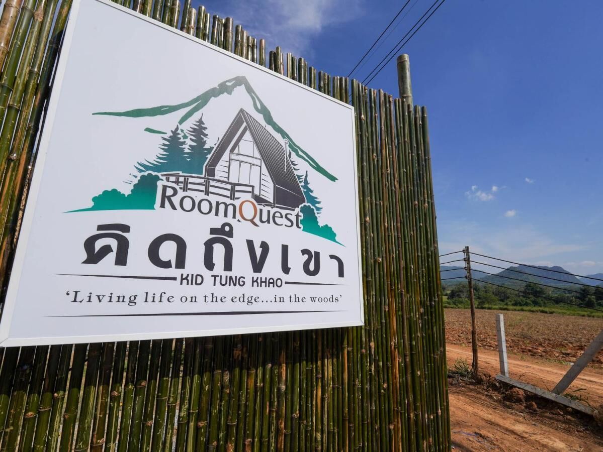 A sign for RoomQuest Mid Tung Khao, featuring a mountain and forest illustration, and the tagline 'Living life on the edge...in the woods'.