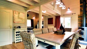 Un coin repas avec une table en bois et des chaises en osier, un buffet blanc où sont rangés des verres à vin, et une cheminée en pierre.