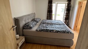 A bedroom with a bed, a nightstand, a wooden floor, and a window with curtains. - Ferienwohnung für Max. 2 Personen (Tacherting)