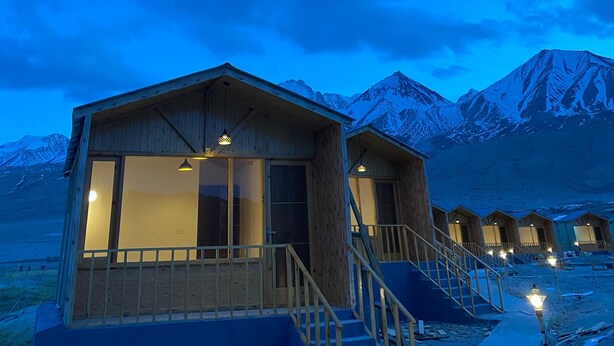 A row of wooden cabins with large windows and a balcony, set against a backdrop of snow-capped mountains under a twilight sky. - Rangjon Resort (Leh)