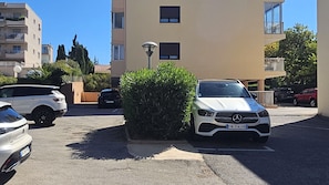 Uma área de estacionamento com um SUV Mercedes-Benz branco, outro SUV branco e um carro preto. Prédios ao fundo e um conjunto de arbustos em primeiro plano.