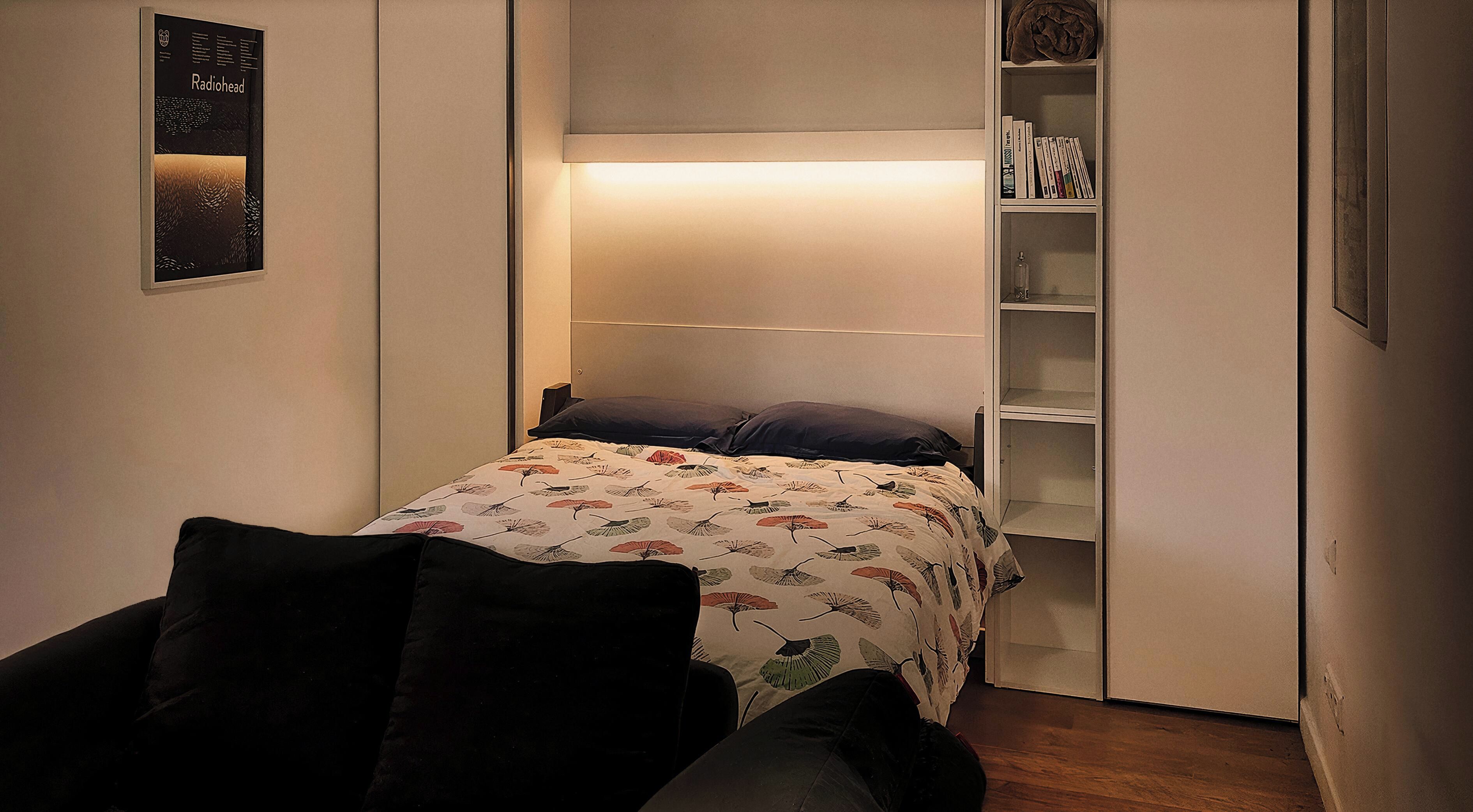 Une chambre à coucher de taille réduite, comprenant un lit, une table de chevet, une étagère avec des livres et une affiche accrochée au mur.