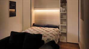 A compact bedroom with a bed, a bedside table, a shelf with books, and a wall-mounted poster.