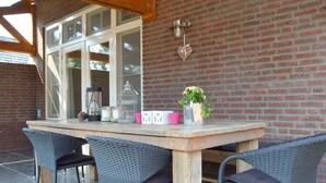 Eine Terrasse im Freien mit einem Holztisch, Korbstühlen, einer Ziegelwand und einem Fenster mit weißen Rahmen.