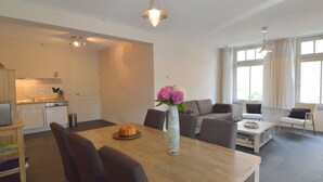 Eine moderne Küche mit weißen Schränken, einem Esstisch mit Stühlen, einer Vase mit Blumen und einem Sofa in einem Wohnbereich mit großen Fenstern.