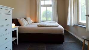 Ein Schlafzimmer mit Bett, Schminktisch, Fenster mit Vorhängen und Nachttisch mit Lampe.