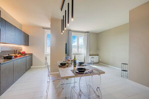 A modern kitchen with a dining area, featuring a central island, clear chairs, and a view of the outdoors through large windows.