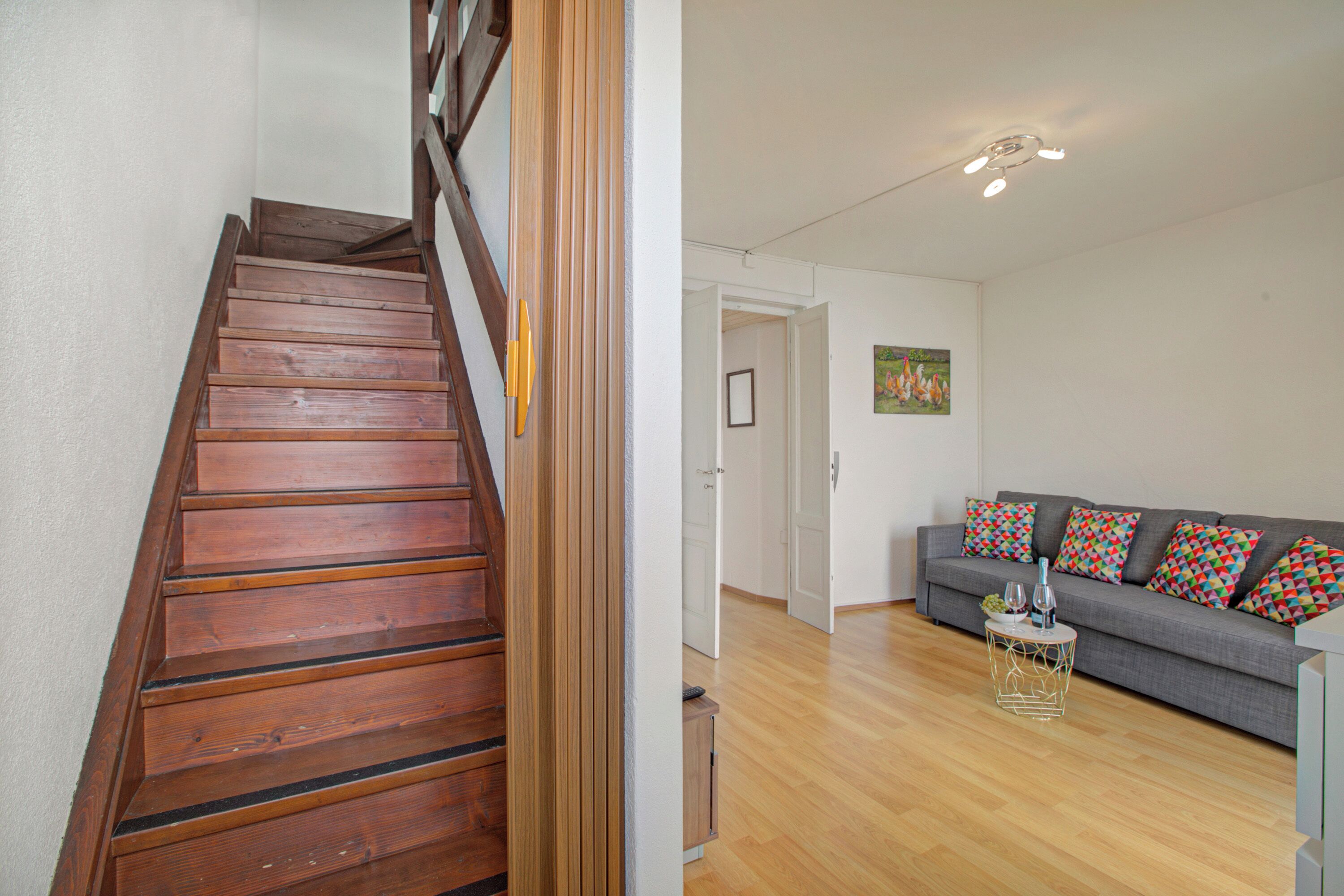 A wooden staircase leading to a room with a grey sofa, a small round table, and a painting on the wall.