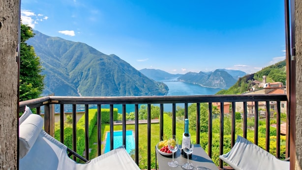 A balcony with a table, a bottle, and a glass of wine, overlooking a mountainous landscape and a body of water.