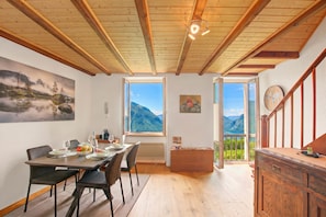 Ein Essbereich mit einem Tisch für vier Personen, Holzbalkendecke, einem Bild an der Wand und Blick auf Berge durch große Fenster.