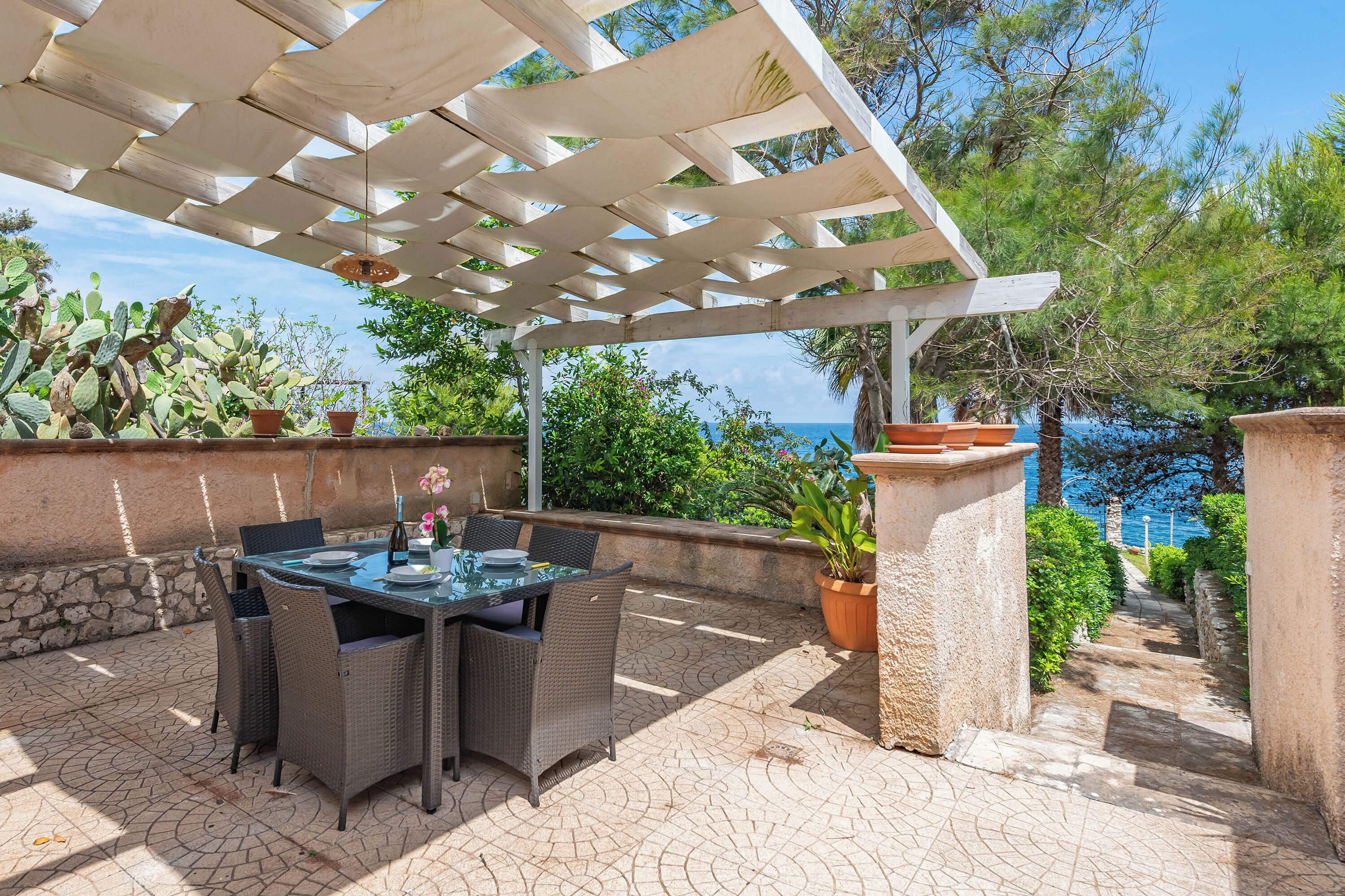 Une terrasse extérieure agrémentée d’une pergola, de meubles en osier, de plantes en pot et offrant une vue sur la mer.