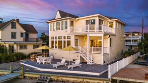 A two-story beachfront house with a balcony, outdoor seating, and a wooden deck.