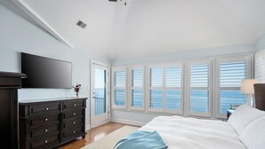 A bedroom with a large bed, a dresser, a flat-screen TV, and a view of the sea through the windows.