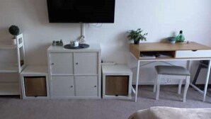 Een knus kamertje met een aan de muur bevestigde televisie, een klein bureau met stoel en een wandrek met kastjes.