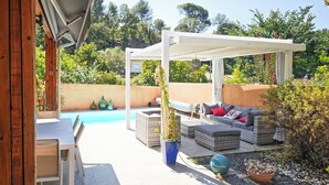 Uma área de estar coberta ao ar livre, com piscina, cercada por vegetação e um pergolado.