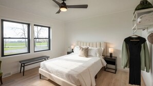 Une chambre à coucher comprenant un lit, deux tables de chevet, un banc, un ventilateur de plafond et une fenêtre donnant sur un parc.