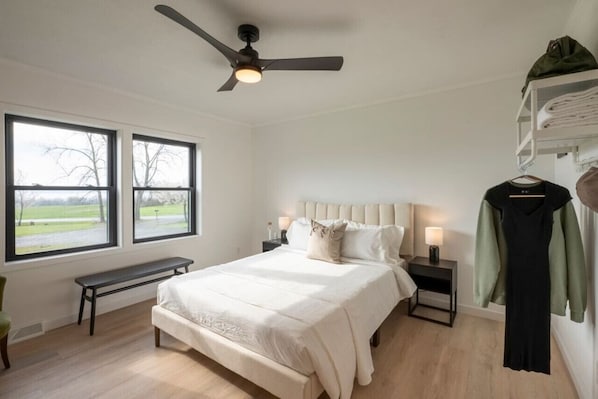 A bedroom with a bed, two bedside tables, a bench, a ceiling fan, and a window with a view of a park.