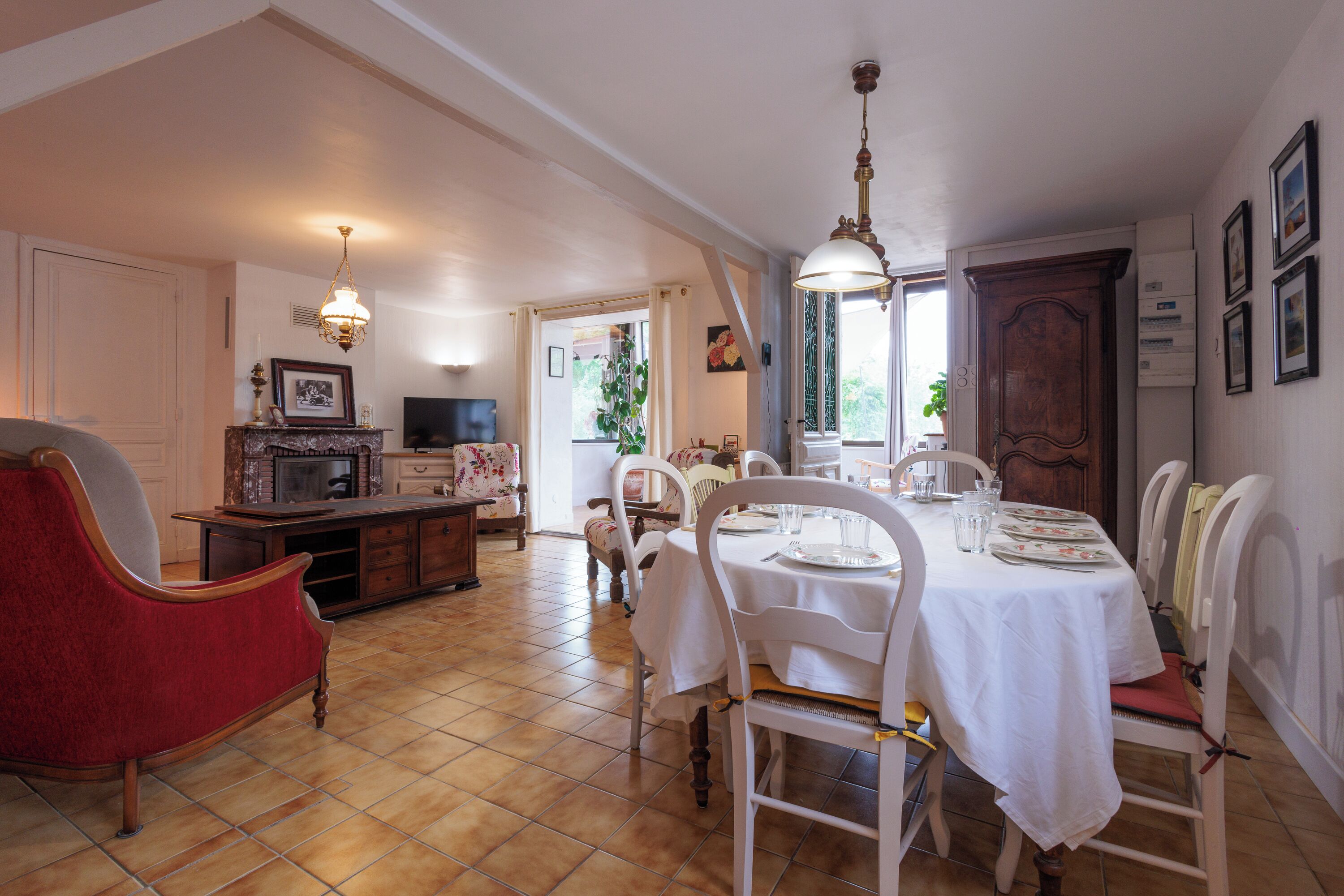 A dining area with a table set for four, surrounded by white chairs. There is a red chair to the left, and a wooden cabinet with a television on top is visible in the background.