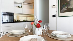 Eine moderne Küche mit weißen Schränken, einem eingebauten Backofen und einem Essbereich, der für eine Mahlzeit gedeckt ist, inklusive weißer Teller, Gläser und einer Vase mit roten Blumen.