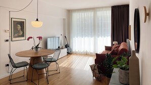 Ein modernes Wohnzimmer mit Esstisch aus Holz, zwei Stühlen, einer Couch und einem großen Fenster mit transparenten Vorhängen.