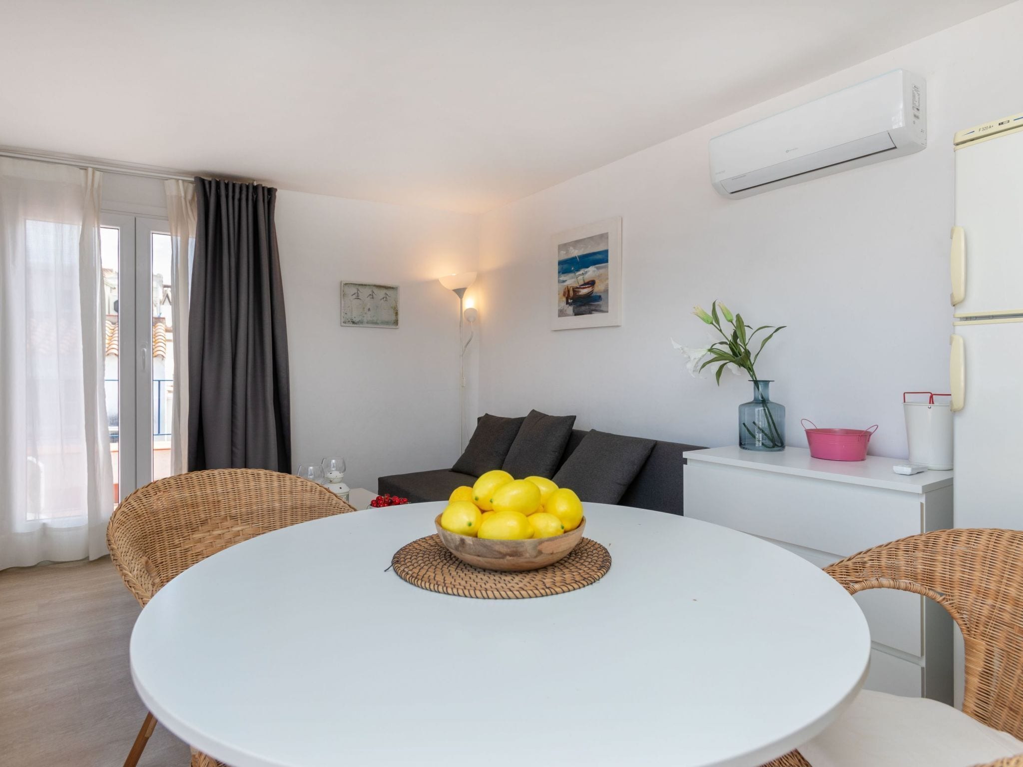 Een moderne woonkamer met een ronde witte tafel, rieten stoelen, een grijze bank en een schaal met citroenen op de tafel.