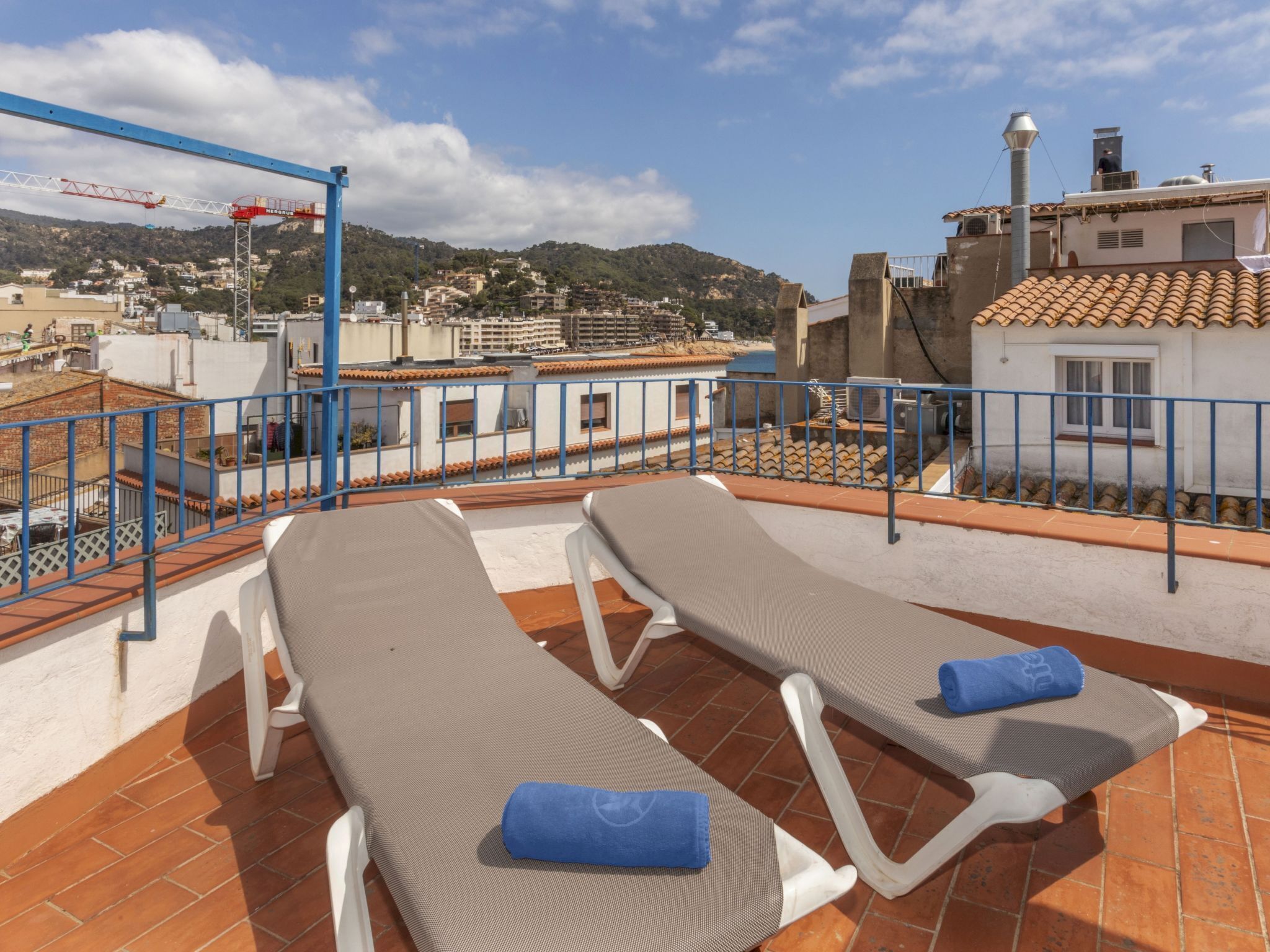 A rooftop terrace with two sun loungers, a blue railing, and a view of the cityscape and hills in the background.