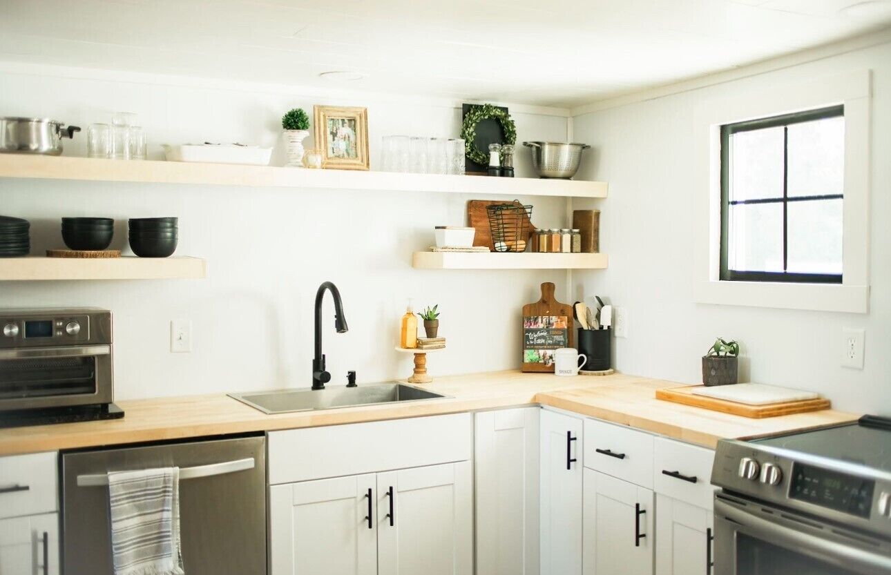 A modern kitchen with white cabinetry, a stainless steel oven, a sink, and various kitchen utensils and decorations on the shelves.