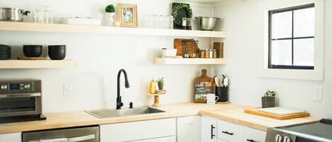A modern kitchen with white cabinetry, a stainless steel oven, a sink, and various kitchen utensils and decorations on the shelves.