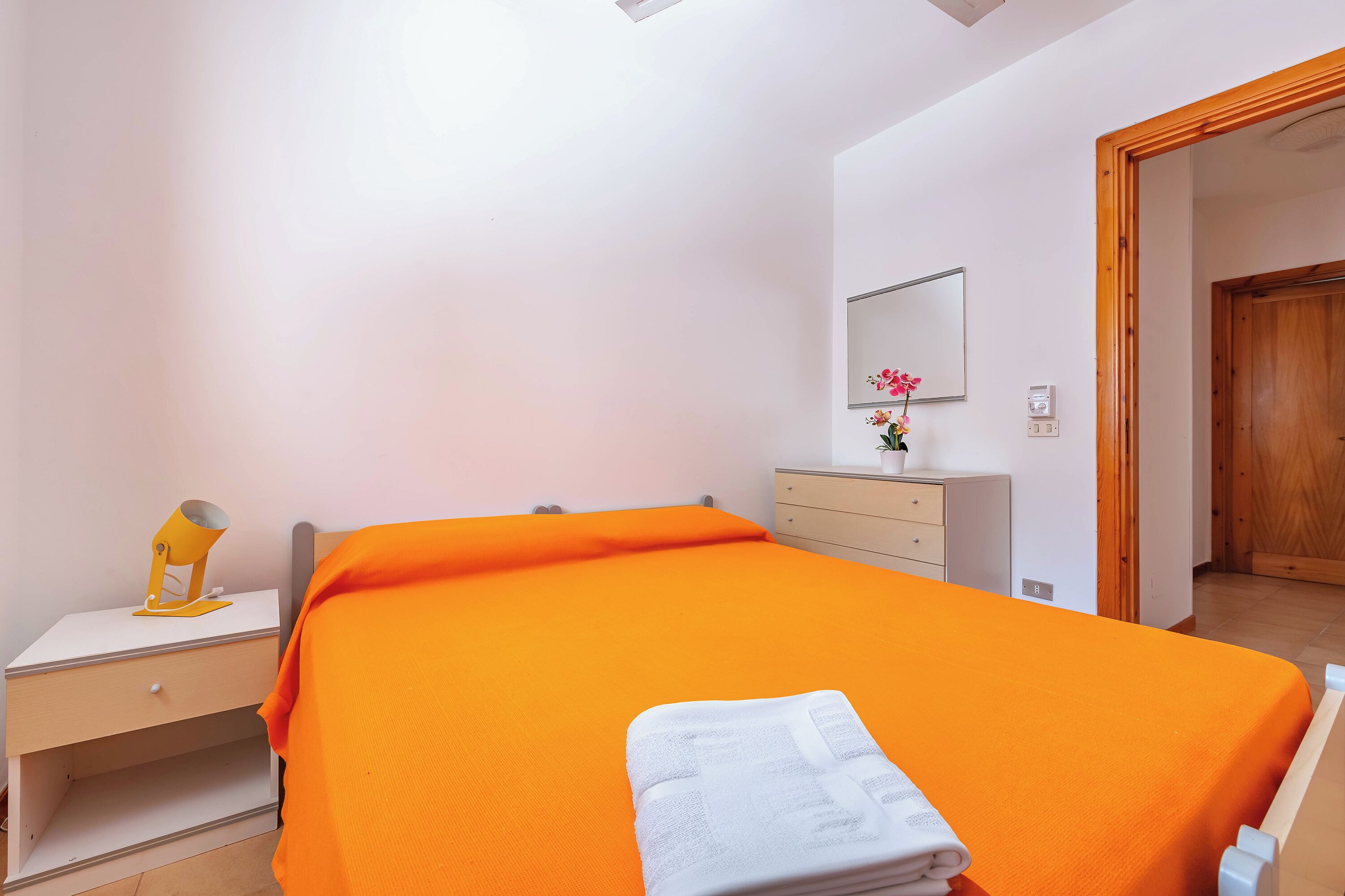 A bedroom with an orange bedspread, a bedside table with a lamp, a dresser, and a mirror on the wall.