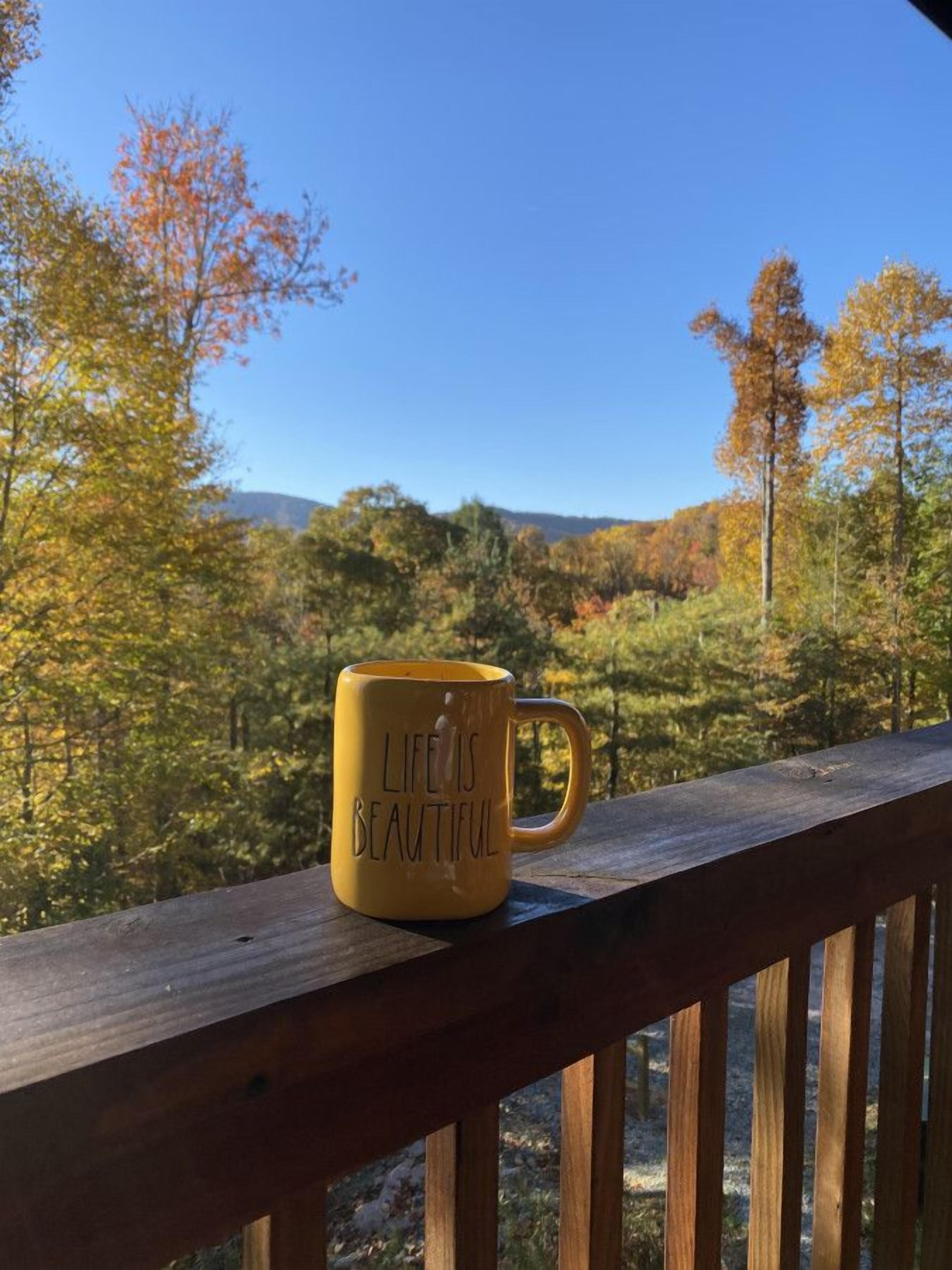 A yellow mug with the inscription "LIFE IS BEAUTIFUL" on a wooden railing with a view of autumn trees and a clear sky.