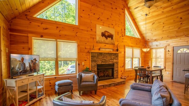 L’intérieur chaleureux d’un chalet avec une cheminée en pierre, des murs en bois et une grande fenêtre donnant sur un paysage verdoyant.