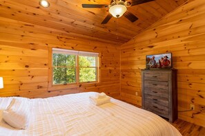 Ein Schlafzimmer einer Holzhütte mit Bett, Kommode, Deckenventilator und Fenster mit Blick auf Bäume.
