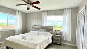 Une chambre à coucher avec un lit, une table de chevet, un ventilateur de plafond et de grandes fenêtres avec des rideaux.