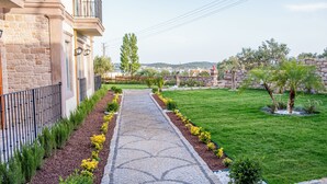 A paved pathway leading to a building with a stone facade, surrounded by a well-maintained garden with various plants and a stone wall in the background. - Cunda Nisa Otel (Ayvalik)