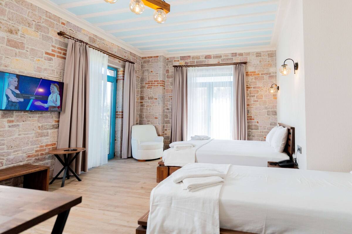 Hotellihuone, jossa on tiiliseinä, kaksi sänkyä, televisio ja puinen penkki.