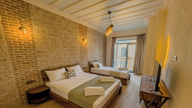 A hotel room with two beds, a wooden bench, a TV, and a balcony with a view. - Cunda Nisa Otel (Ayvalik)