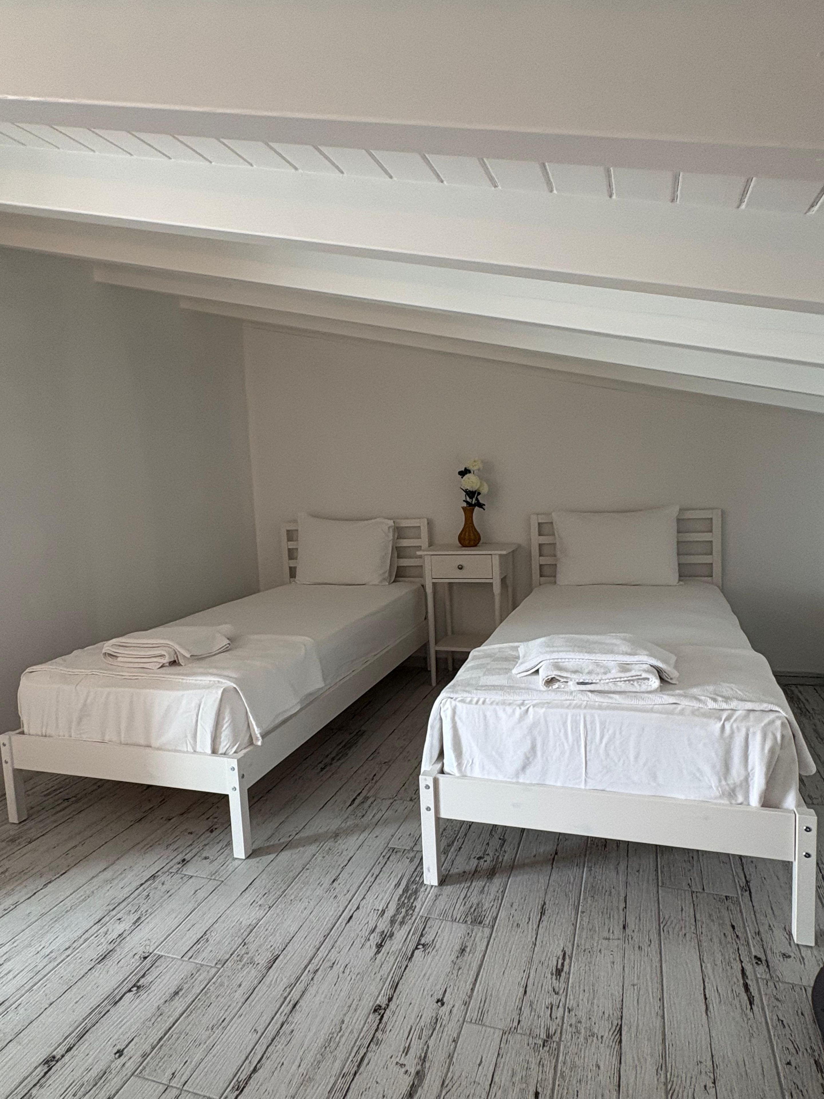 A room with two single beds, a small bedside table with a vase, and a wooden floor.