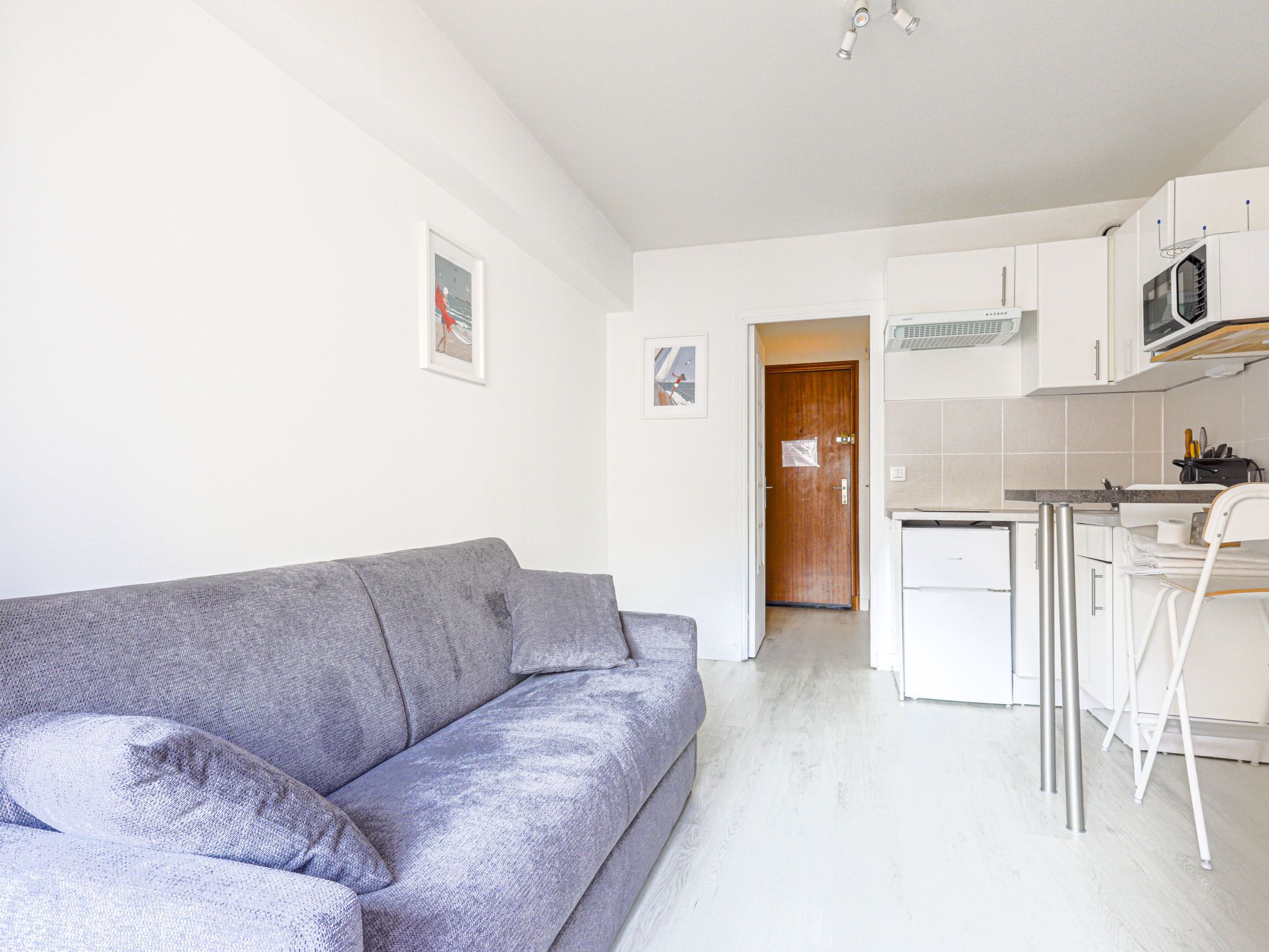 Un espace de vie compact comprenant un canapé gris, une kitchenette avec des meubles blancs et une porte en bois donnant sur une autre pièce.