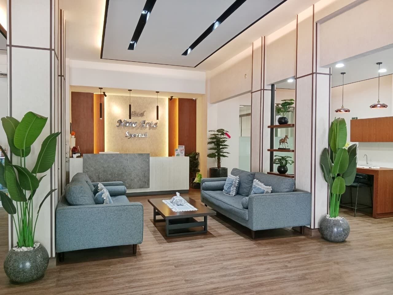 A modern hotel lobby with a reception desk, seating area, and a kitchen visible through a glass partition.