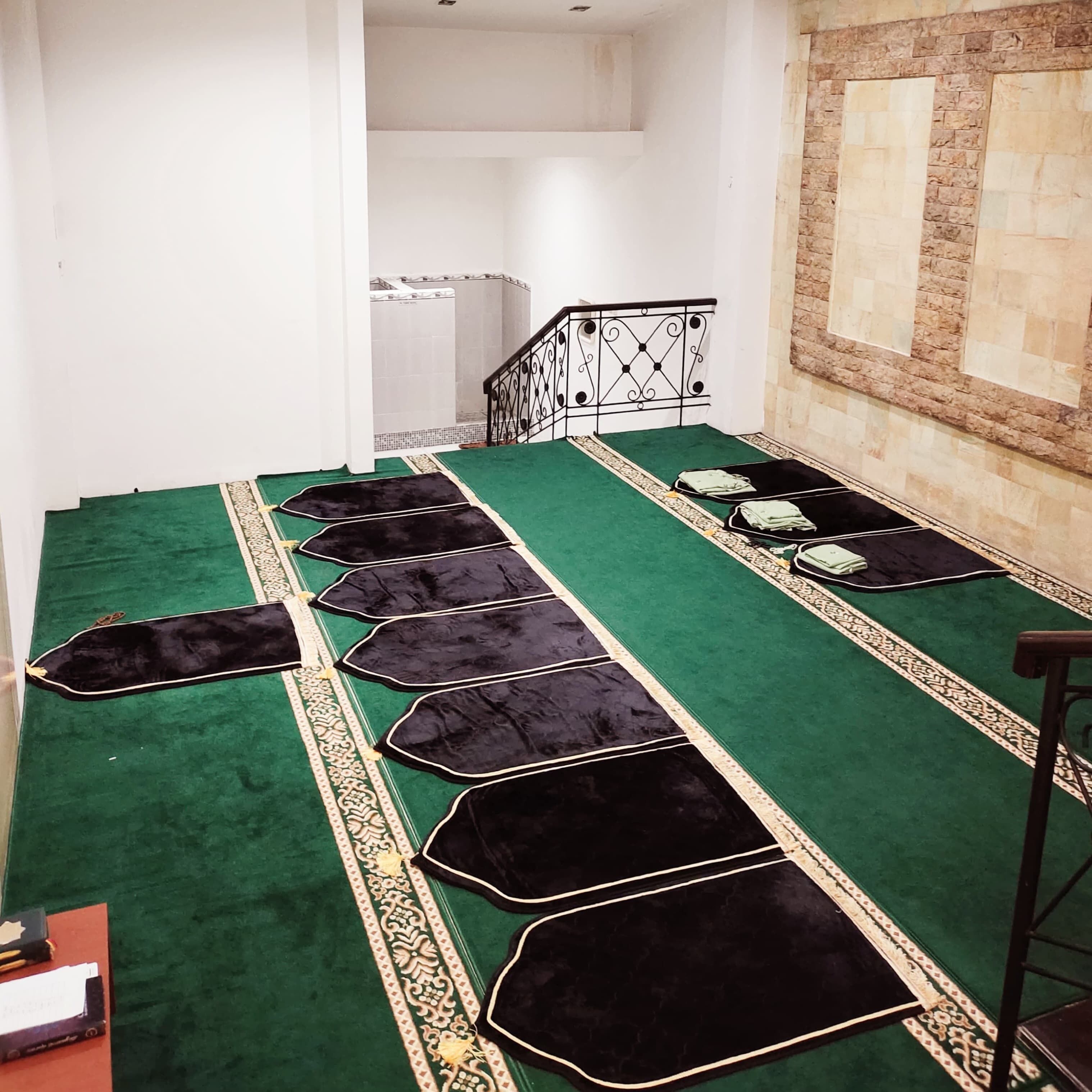 A prayer area with a green carpet, black prayer mats, and a staircase with a metal railing.